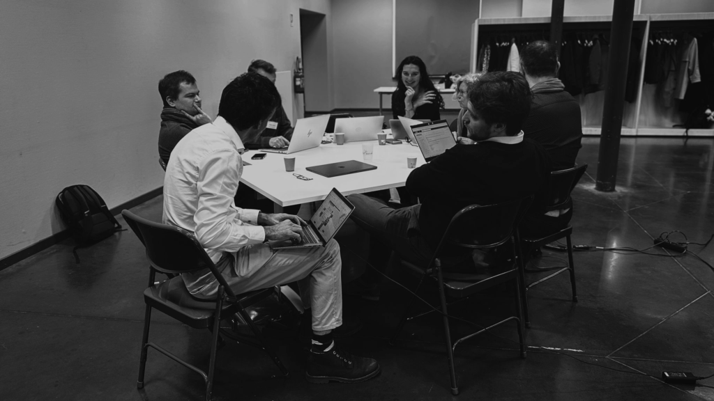 Photo en noir et blanc d’une réunion de travail avec plusieurs participants assis autour d’une table ronde. Les participants, certains avec des ordinateurs portables ouverts devant eux, semblent discuter et collaborer activement. L’ambiance est professionnelle et dynamique, reflétant l’engagement des acteurs pour un tourisme inclusif et innovant. En arrière-plan, on distingue une salle moderne avec des colonnes et des murs en bois clair.