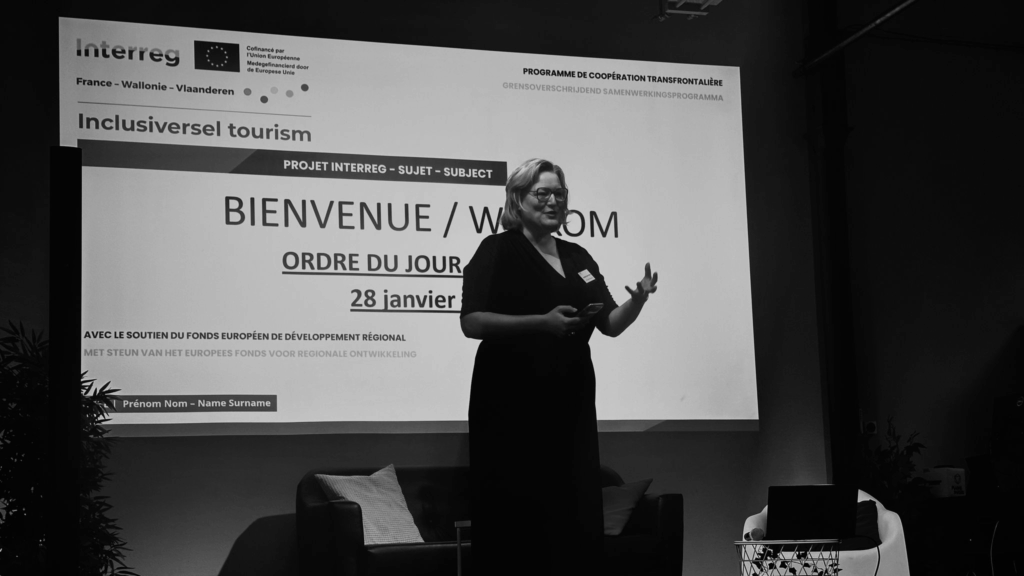 Photo en noir et blanc d’une intervenante debout devant un écran géant lors d’un événement dédié au projet Inclusiversel tourism. L’écran affiche le titre 'BIENVENUE / WELKOM' suivi de 'ORDRE DU JOUR' et de la date '28 janvier'. En haut de l’écran, on voit les logos d’Interreg, de l’Union Européenne, et la mention 'France - Wallonie - Vlaanderen'. L’intervenante, tenant un micro, semble animer une présentation devant un public attentif. L’ambiance est professionnelle et collaborative, typique des ateliers transfrontaliers.