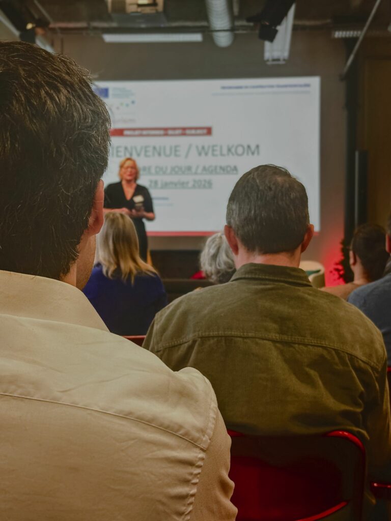 Photo d’une présentation publique avec un public assis face à une intervenante debout devant un écran. L’écran affiche le titre 'BIENVENUE / WELKOM' suivi de 'ORDRE DU JOUR / AGENDA' et de la date '28 janvier 2026'. Les logos d’Interreg et de l’Union Européenne sont visibles en haut de l’écran. L’intervenante, tenant un micro, semble expliquer les objectifs du projet Inclusiversel tourism à un public attentif. L’ambiance est professionnelle et engageante, typique des événements de lancement ou de présentation de projets transfrontaliers.