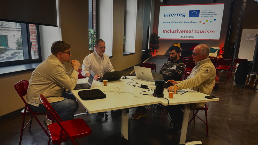 Photo d’un atelier de travail en groupe avec quatre participants assis autour d’une table rectangulaire. Chaque personne utilise un ordinateur portable, et des gobelets en carton sont disposés sur la table, indiquant une pause café ou une session de travail informelle. En arrière-plan, une affiche murale affiche les logos d’Interreg, de l’Union Européenne, et le titre 'Inclusiversel tourism' avec la date '28.01.2026'. La salle est lumineuse grâce à de grandes fenêtres, et des plantes vertes ajoutent une touche chaleureuse à l’espace.