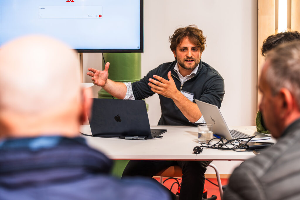 Ruben Dedecker, Doctorant (Ghent University) en pull noir anime un atelier collaboratif autour d'une table, avec des participants attentifs et des ordinateurs portables ouverts.