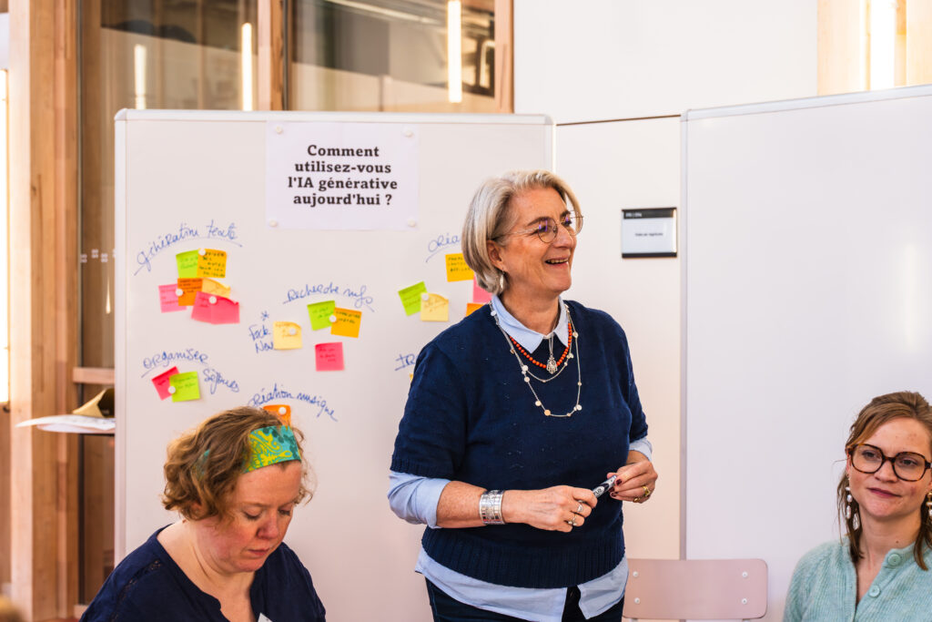 France Lefebvre du Preÿ, Responsable pédagogique, en pull bleu anime un atelier devant un tableau blanc couvert de post-it colorés, avec la question "Comment utilisez-vous l'IA générative aujourd'hui ?" écrite au centre.