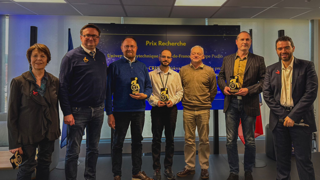 Photo de groupe lors de la remise des Prix Recherche dans le cadre des Prix de l’Excellence Handi Tech. Sept personnes, debout devant un écran affichant le texte 'Prix Recherche' et les logos des partenaires, tiennent fièrement leurs trophées dorés en forme de silhouette en fauteuil roulant. Les lauréats, vêtus de manière formelle ou décontractée, sourient à la caméra, symbolisant leur fierté et leur engagement pour l’innovation dans le domaine de l’accessibilité. En arrière-plan, on distingue des drapeaux français et européens, soulignant le caractère officiel et transfrontalier de l’événement.