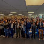 Photo de groupe prise lors de la cérémonie de remise des Prix de l’Excellence Handi Tech 2026 à la French Tech Lille. Un grand nombre de participants, debout dans une salle moderne avec des chaises bleues et un écran en arrière-plan, lèvent les pouces en signe de célébration. Plusieurs personnes au premier rang tiennent des trophées dorés en forme de silhouette en fauteuil roulant, symbolisant leur reconnaissance dans le domaine de l’innovation et de l’accessibilité. L’ambiance est joyeuse et collaborative, reflétant l’engagement des lauréats et des acteurs pour un tourisme et une technologie inclusifs.