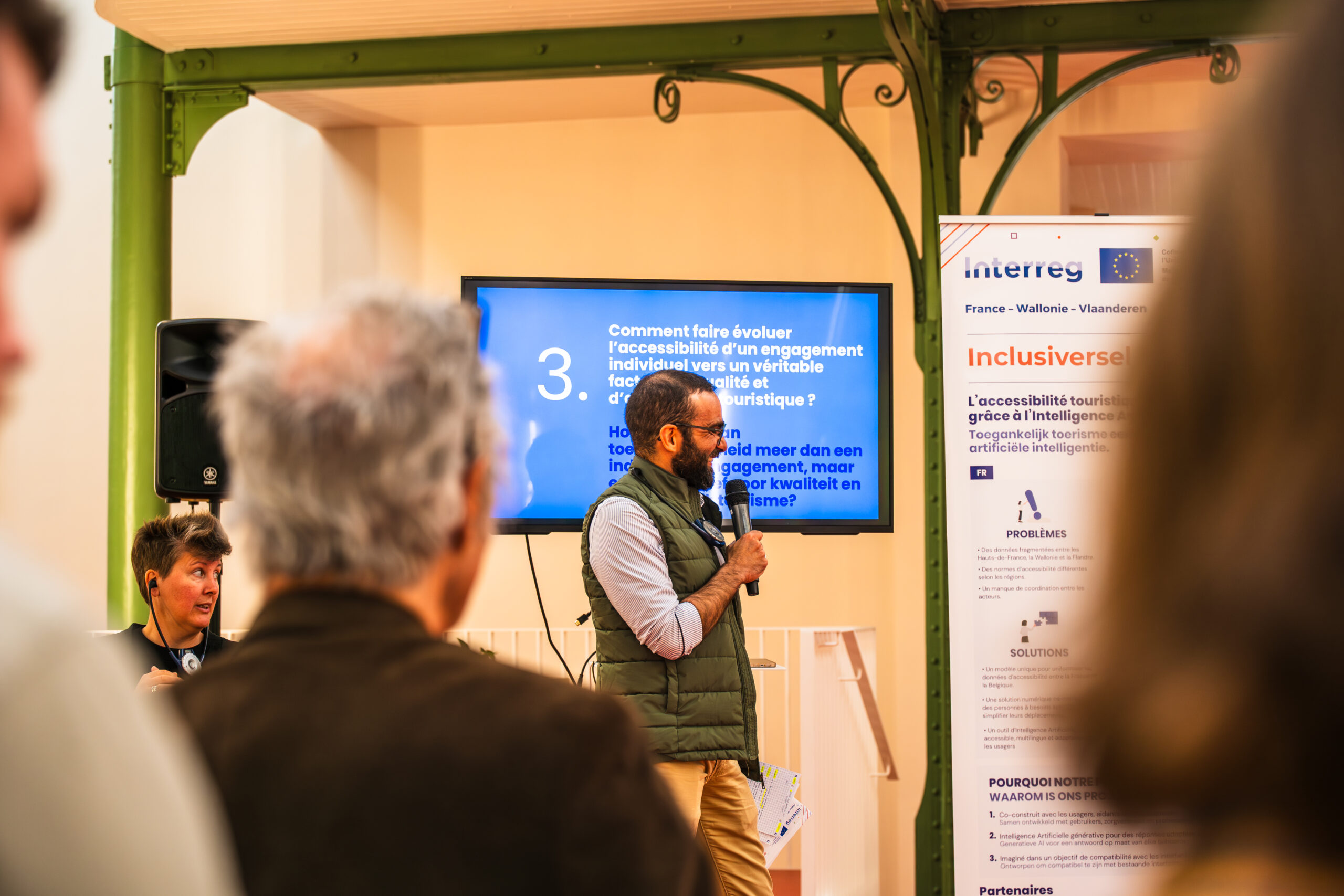 Photo de Daniel Grancho, Responsable Partenariats (Picto Access), en train de parler lors d’une présentation sur l’accessibilité et l’intelligence artificielle, dans le cadre du projet Inclusiversel Tourism. L’orateur, debout devant un écran affichant des questions en français et en néerlandais, tient un micro et semble animer une discussion. À gauche, une participante écoute attentivement, tandis qu’en arrière-plan, une bannière Interreg France-Wallonie-Vlaanderen et une affiche du projet Inclusiversel Tourism sont visibles. La salle, avec ses murs en bois clair et ses structures métalliques vertes, reflète une ambiance professionnelle et collaborative, typique des ateliers transfrontaliers."