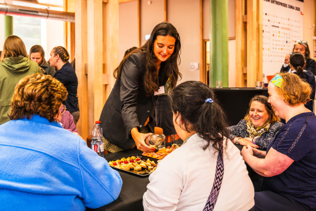 Photo prise lors de notre événement de lancement en mai 2026. Ambre Santer, Coordinatrice du projet Interreg Inclusiversel tourism, sert des amuse-bouches à un groupe de participants assis autour d’une table. Les convives, variés en âge et en apparence, discutent et partagent un moment de convivialité. En arrière-plan, d’autres participants interagissent dans une salle lumineuse aux murs en bois clair, avec des affiches et des éléments décoratifs. Une bouteille d’eau et des verres sont visibles sur la table, renforçant l’ambiance chaleureuse et collaborative de l’événement.