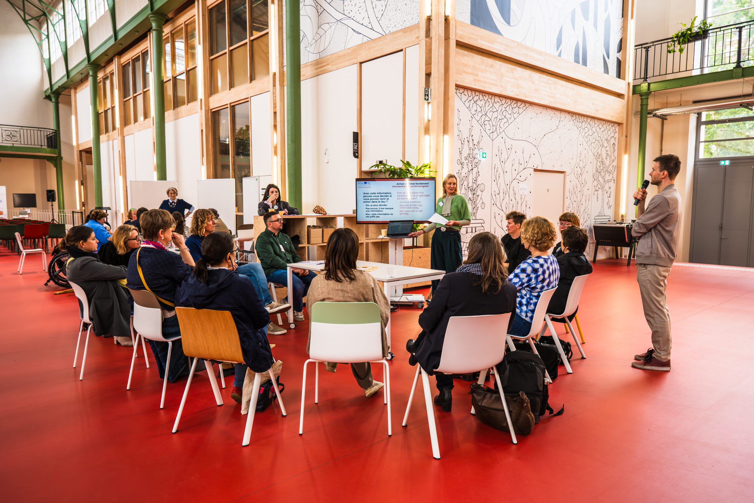 Photo d’un atelier collaboratif dans un espace moderne et lumineux, avec de grandes fenêtres et des colonnes en bois. Un groupe de participants, assis en cercle sur des chaises blanches et colorées, écoute deux animateurs debout. L’un des animateurs, tenant un micro, semble expliquer un point affiché sur un écran derrière lui. Une table centrale supporte des documents et un ordinateur portable. L’ambiance est dynamique et inclusive, illustrant la co-construction des solutions pour le projet Inclusiversel Tourism. Les murs décorés de motifs artistiques et les plantes vertes ajoutent une touche chaleureuse à l’espace.