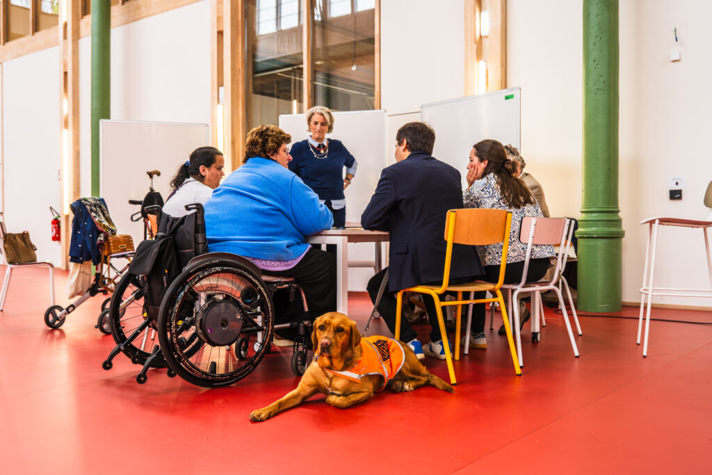 Photo d’un atelier collaboratif dans le cadre du projet Inclusiversel Tourism. Plusieurs participants, dont une personne en fauteuil roulant, sont assis autour d’une table en discussion. Un chien d’assistance, portant un gilet orange, est allongé au premier plan, symbolisant l’inclusivité et le soutien aux personnes en situation de handicap. En arrière-plan, une femme debout anime l’échange, tandis que d’autres participants, assis sur des chaises colorées, écoutent attentivement. La salle, avec ses colonnes vertes et son sol rouge, offre un cadre moderne et accessible, reflétant l’engagement du projet pour un tourisme inclusif et adapté à tous.