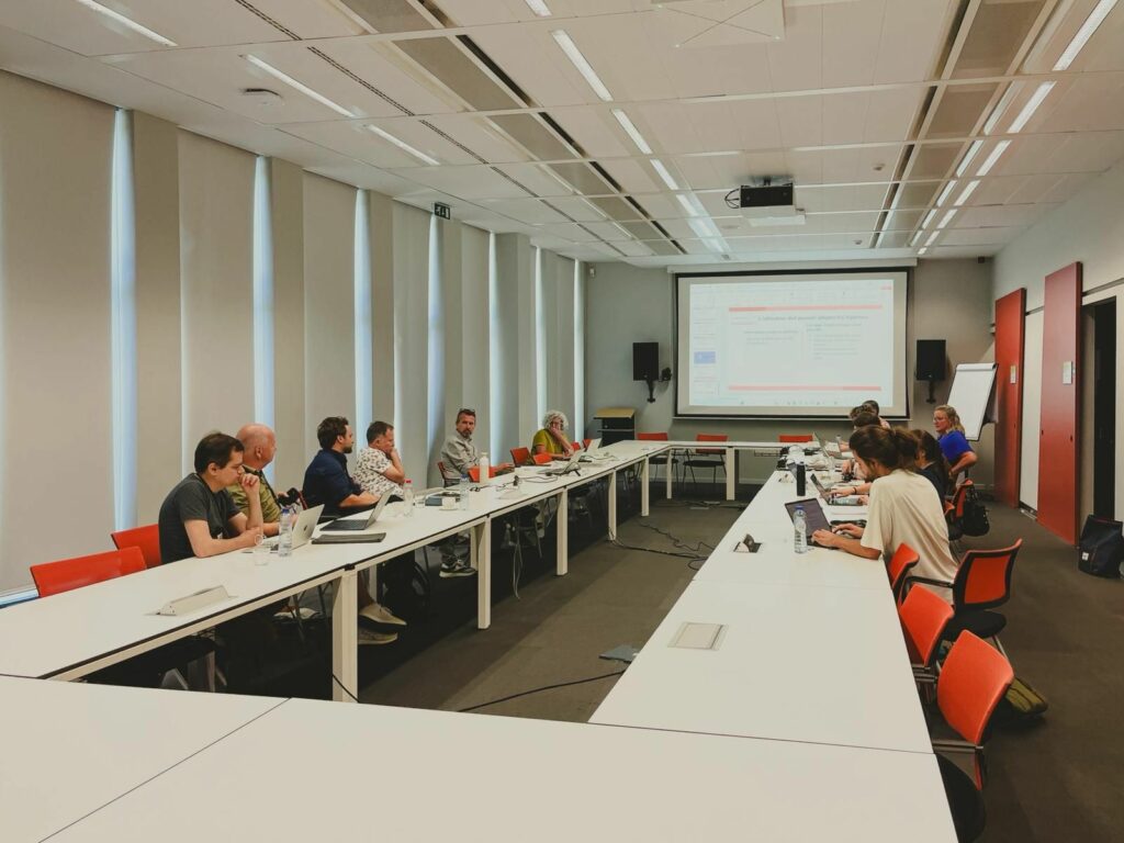 Une photo en grand angle montre une douzaine de personnes assises autour d'une longue table blanche en U dans une salle de conférence moderne et lumineuse. Elles écoutent une présentation projetée sur un grand écran au fond de la pièce. Certains participants ont des ordinateurs portables ouverts devant eux. Des stores verticaux blancs couvrent les fenêtres sur le mur de gauche. Des chaises orange vif contrastent avec le mobilier de bureau.