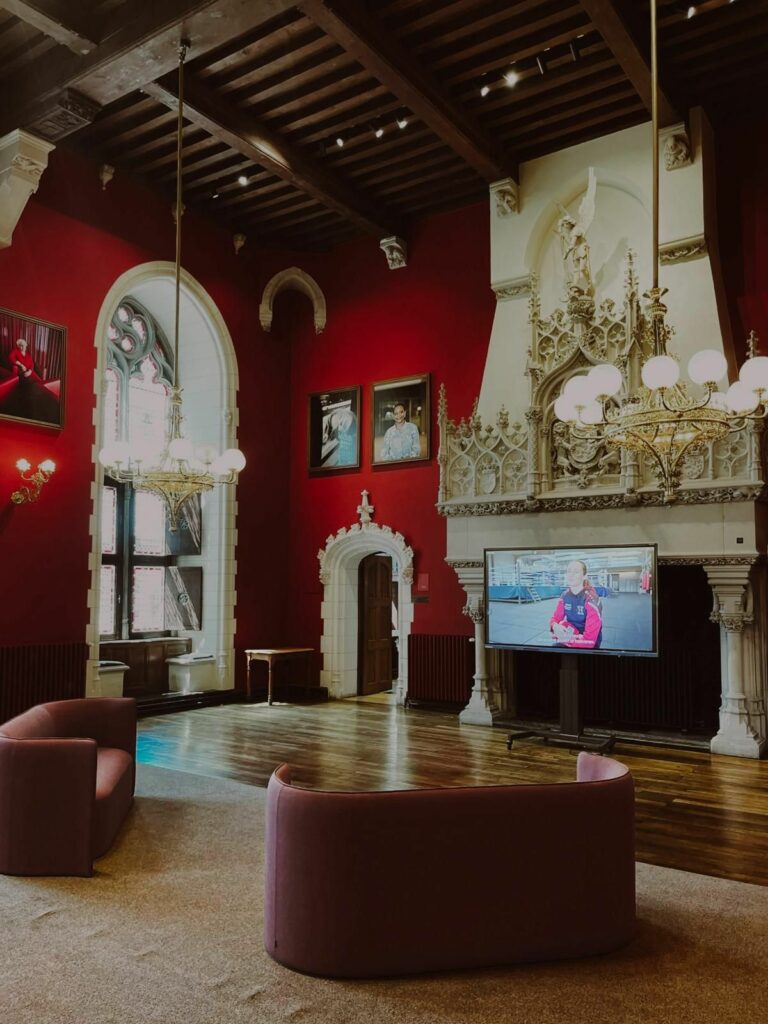 Vue d'un salon luxueux à l'architecture néo-gothique au Provinciaal Hof de Bruges. La pièce est spectaculaire avec ses murs rouge vif, son plafond en bois sombre à poutres apparentes et ses lustres complexes. Une grande cheminée en pierre sculptée orne le mur du fond, à côté d'un écran de télévision noir affichant une vidéo. Deux canapés incurvés en velours rose sont disposés au premier plan sur un tapis beige, face à la cheminée. Des fenêtres gothiques en ogive laissent entrer la lumière naturelle sur la gauche.