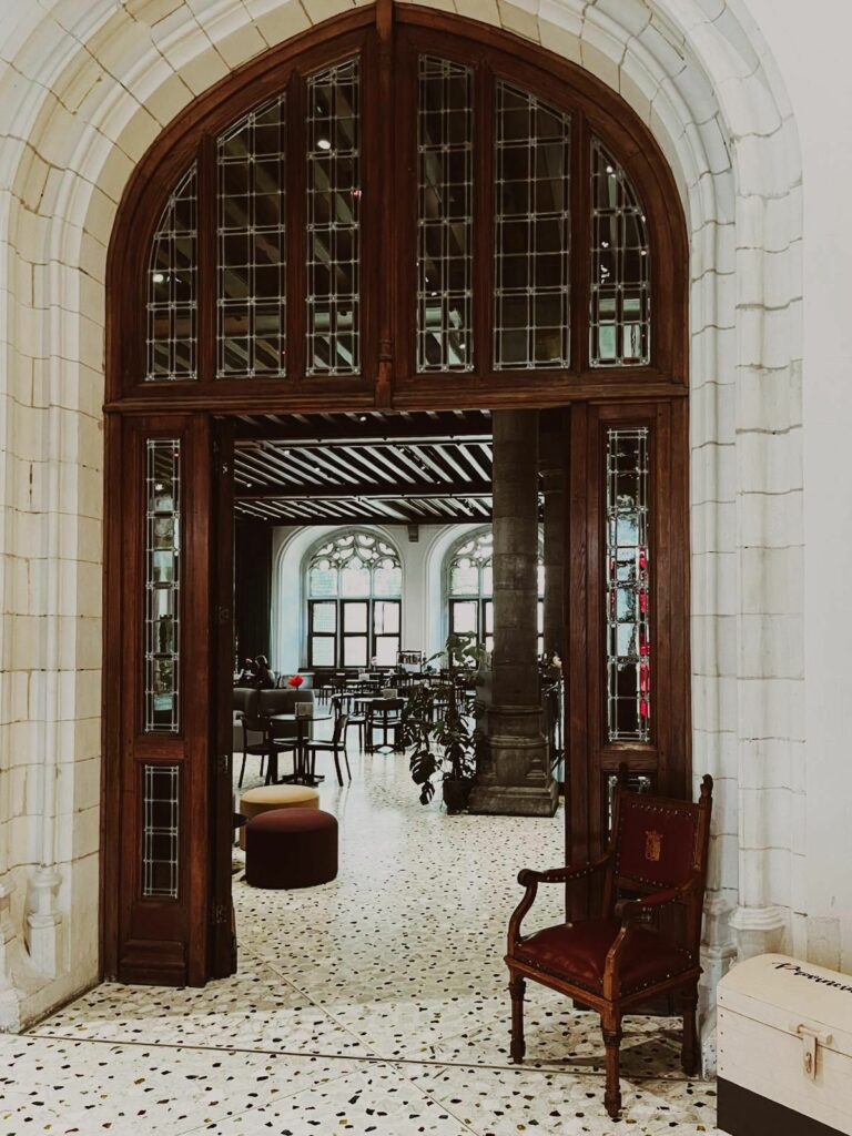 Une grande porte voûtée en bois sombre et vitraux s'ouvre sur un espace baigné de lumière. Au premier plan, un fauteuil en cuir rouge et des poufs sont posés sur un sol en terrazzo. En arrière-plan, à travers les arches en pierre, on aperçoit la salle du Hofcafé installée sous de hautes fenêtres gothiques.