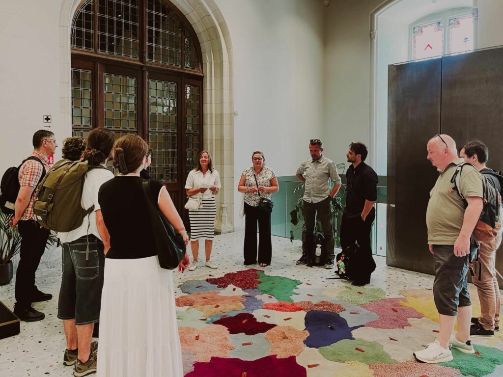 Un groupe d'une dizaine de personnes écoute deux femmes qui se tiennent debout devant eux sur un tapis coloré dans une grande salle d'exposition au Provinciaal Hof. Marieke Vanloocke, l'une des deux femmes, s'adresse au groupe. La plupart des participants ont le dos tourné à la caméra et portent des sacs à dos. La salle est vaste, avec des murs blancs et une grande fenêtre gothique en arrière-plan.