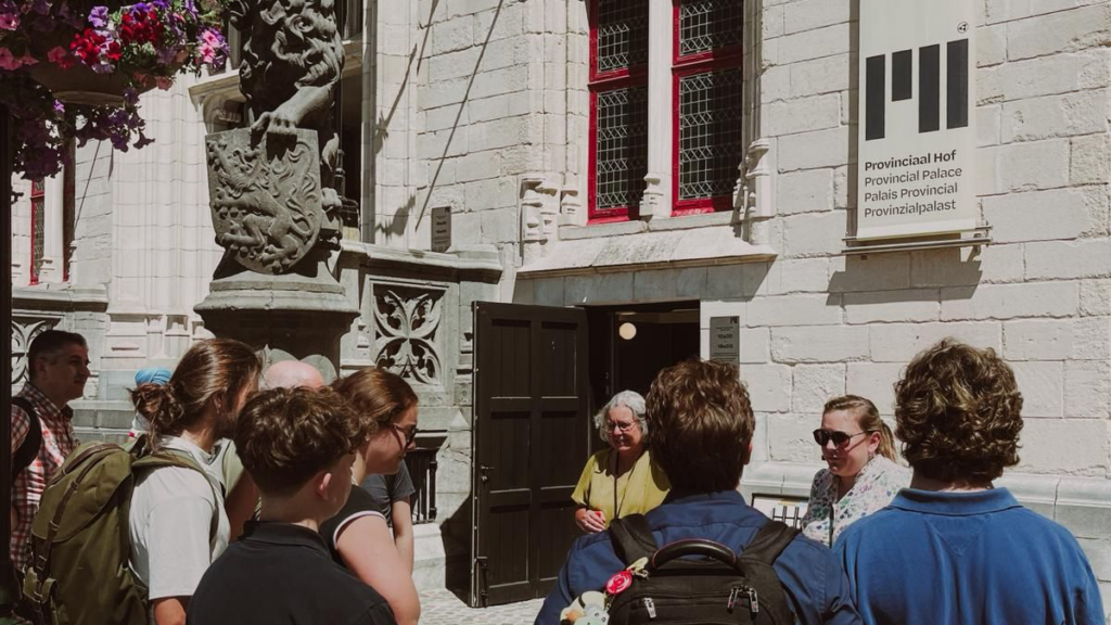 Un groupe de sept personnes, vu de dos, marche vers l'entrée du Provinciaal Hof de Bruges par une journée ensoleillée. La façade en pierre de style néo-gothique du bâtiment est ornée de détails complexes. Une statue de lion portant un blason se dresse à gauche, avec des pots de pétunias roses suspendus au-dessus. Une bannière blanche avec des rayures rouges et roses et le logo "PH" pend sur la droite. Les visiteurs portent des sacs à dos et des t-shirts.