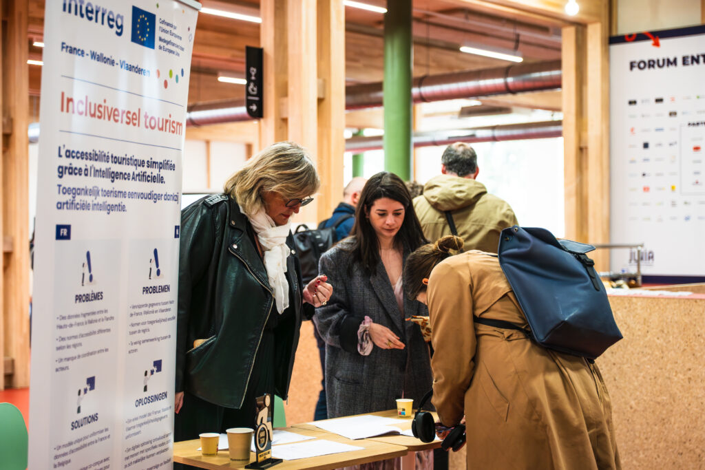 Trois participantes réunies autour d'une table d'accueil devant la bannière bilingue du projet Inclusiversel tourism.